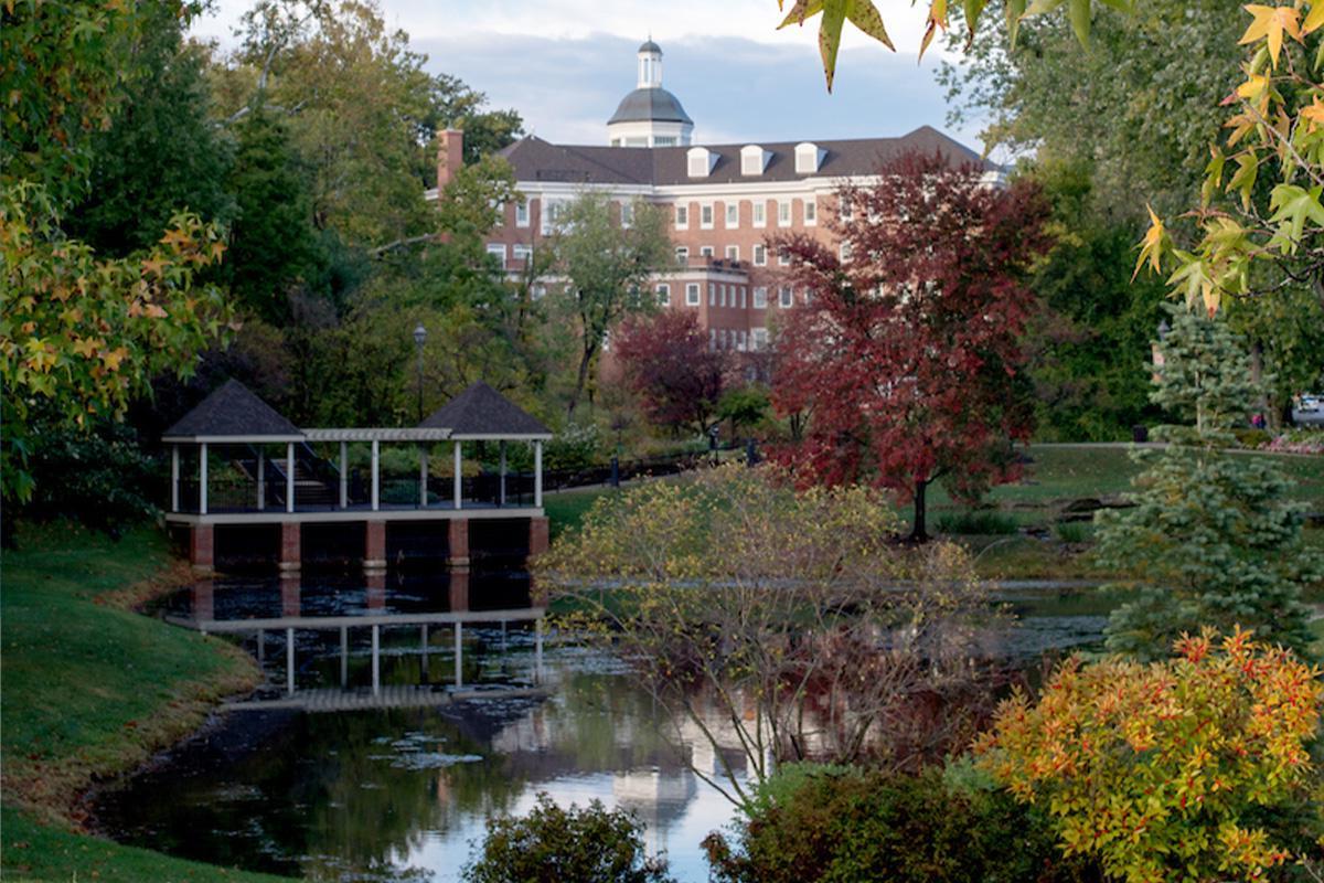 Emeriti Park at Ohio University, featuring fall foliage surrounding a small pond on campus, with a brick building in the background